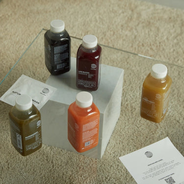 Vier Flaschen DALUMA 1 TAG SAFTFASTEN RESET stehen auf einem quadratischen weißen Block auf einem beigen Teppich. In der Nähe befinden sich zwei weiße Papiere mit Text. Jede Flasche hat einen weißen Verschluss und ein Etikett - ideal für Ihre nächste Saftkur oder Saftfasten.