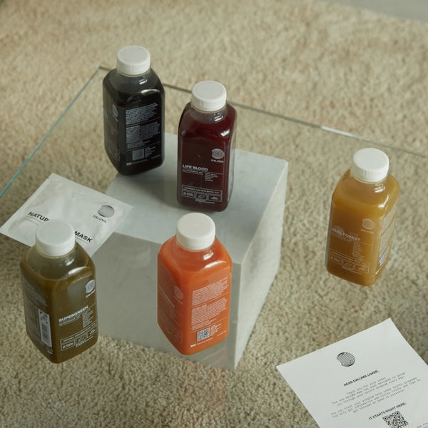 Vier Flaschen DALUMA 1 TAG SAFTFASTEN RESET stehen auf einem quadratischen weißen Block auf einem beigen Teppich. In der Nähe befinden sich zwei weiße Papiere mit Text. Jede Flasche hat einen weißen Verschluss und ein Etikett - ideal für Ihre nächste Saftkur oder Saftfasten.