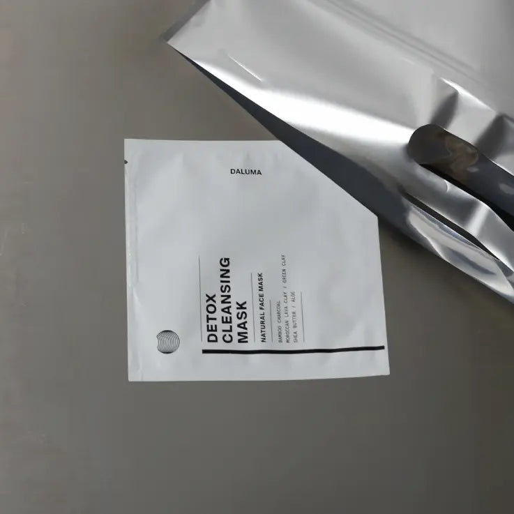 Eine weiße Packung mit der Aufschrift Detox Cleansing Mask liegt auf einer grauen Fläche neben einem teilweise gefalteten Blatt aus reflektierender Silberfolie. Die Packung hat einen minimalen schwarzen Text und ein klares Design.