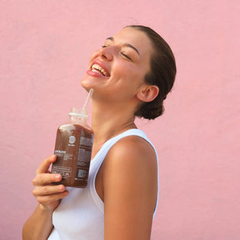 Eine lächelnde Frau in einem weißen Tank-Top hält eine Flasche Schokoladengetränk mit einem Strohhalm und steht vor einem rosa Hintergrund. Sie sieht glücklich aus und neigt ihren Kopf leicht zurück, um den Moment zu genießen.