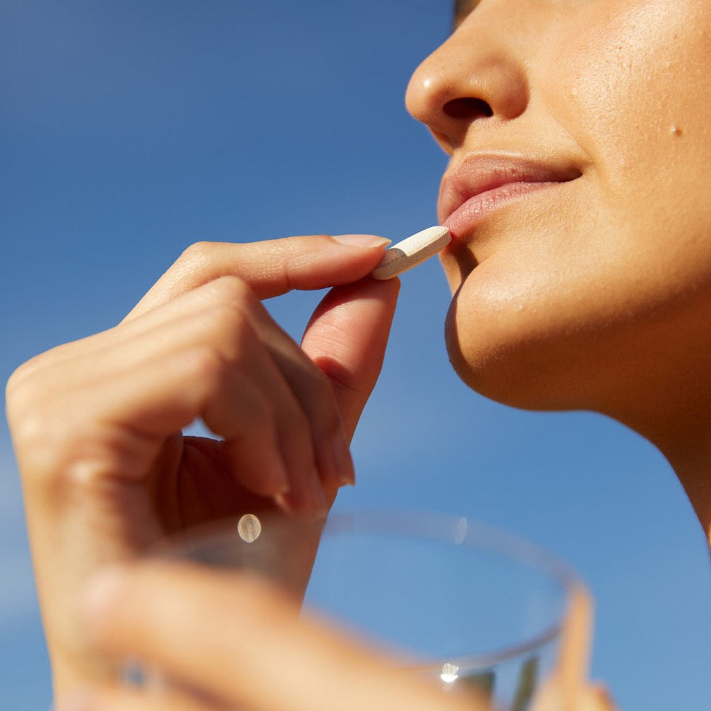 Nahaufnahme von Gesicht und Händen einer Person, die eine FACE GLOW DUO-Kapsel von DALUMA an die Lippen hält, mit einem Glas Wasser in der anderen Hand und einem klaren blauen Himmel im Hintergrund.