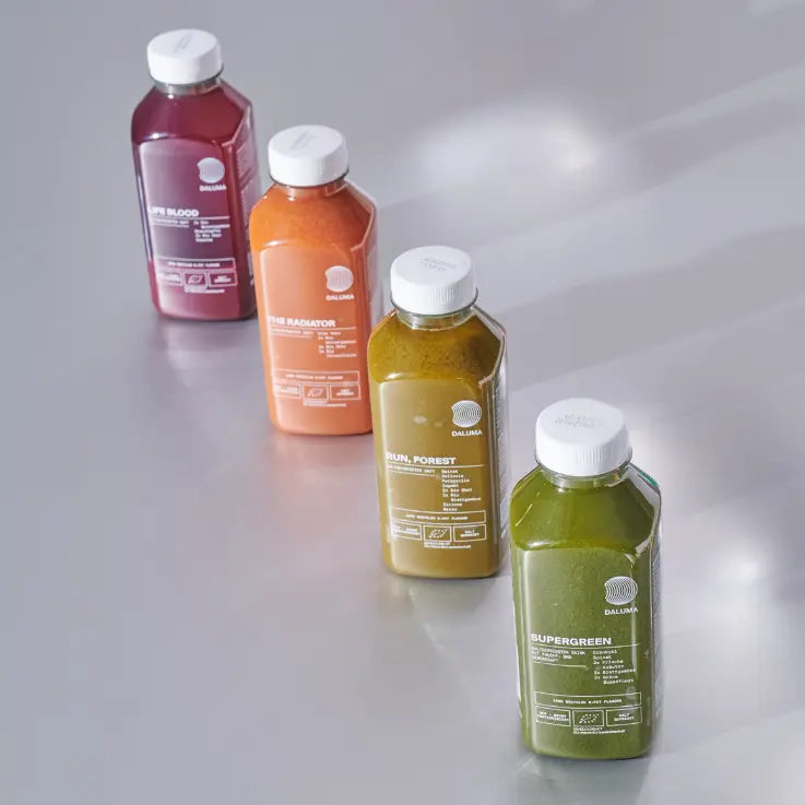 Vier Flaschen mit kaltgepresstem Saft in verschiedenen Farben (lila, orange, gelb-grün und grün) sind diagonal auf einer hellen Fläche angeordnet und jeweils mit eindeutigen Namen und weißen Verschlüssen versehen.