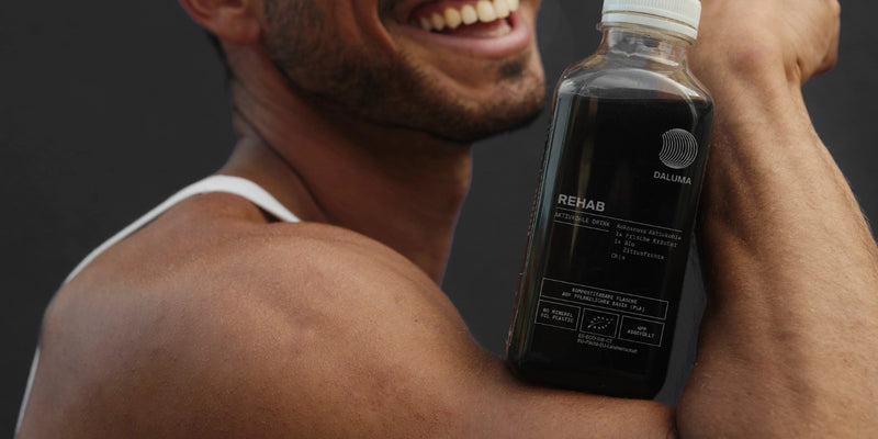 Eine lächelnde Person in einem weißen Tank-Top hält eine Flasche mit der Aufschrift REHAB dicht an ihre Schulter und präsentiert das Getränk. Die Flasche ist dunkel und weist Branding und Produktdetails auf dem Etikett auf.