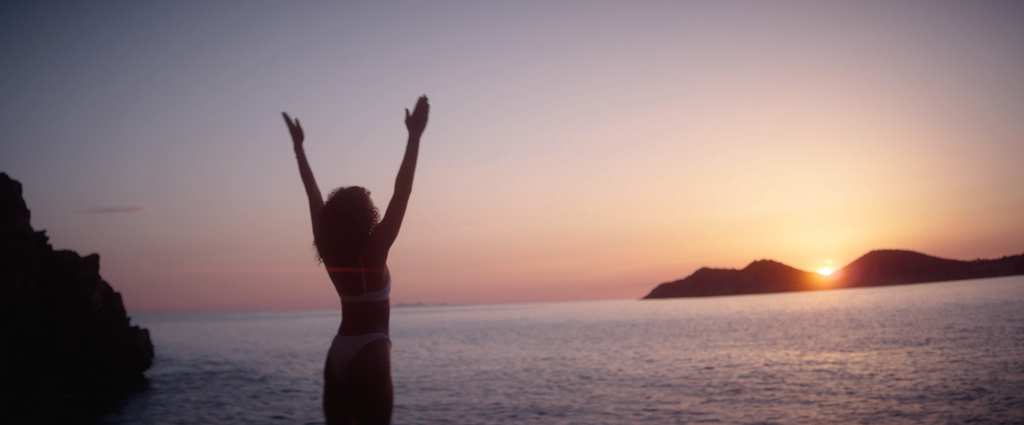 Frau im Bikini steht mit erhobenen Armen und blickt auf ein ruhiges Meer bei Sonnenuntergang. Im Hintergrund sind schemenhafte Berge und ein bunter Himmel zu sehen, die eine friedliche, fröhliche Szene schaffen.