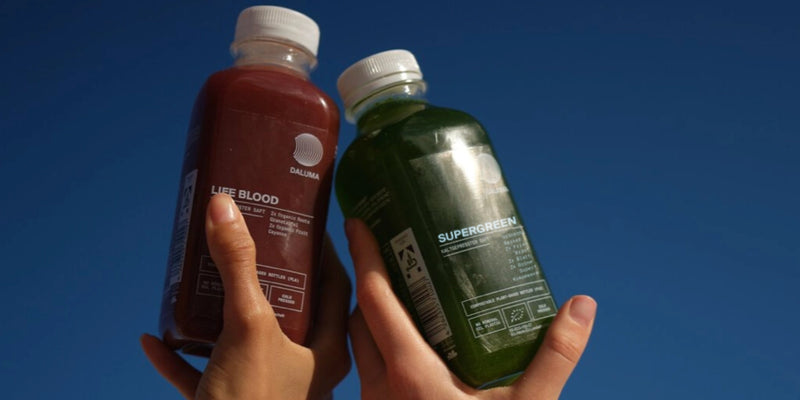 Zwei Hände halten Saftflaschen gegen einen klaren blauen Himmel - eine Flasche enthält einen roten Saft mit der Aufschrift LIFE BLOOD, die andere einen grünen Saft mit der Aufschrift SUPERGREEN.