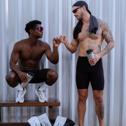 Zwei sportliche Männer mit Sonnenbrillen, schwarzen Shorts und Shakerflaschen in der Hand stoßen sich mit den Fäusten aneinander. Einer sitzt auf einer Bank, der andere steht mit einem Handtuch auf dem Kopf. Sie stehen vor einer Metallwand mit Sporttaschen und Paketen auf dem Boden.