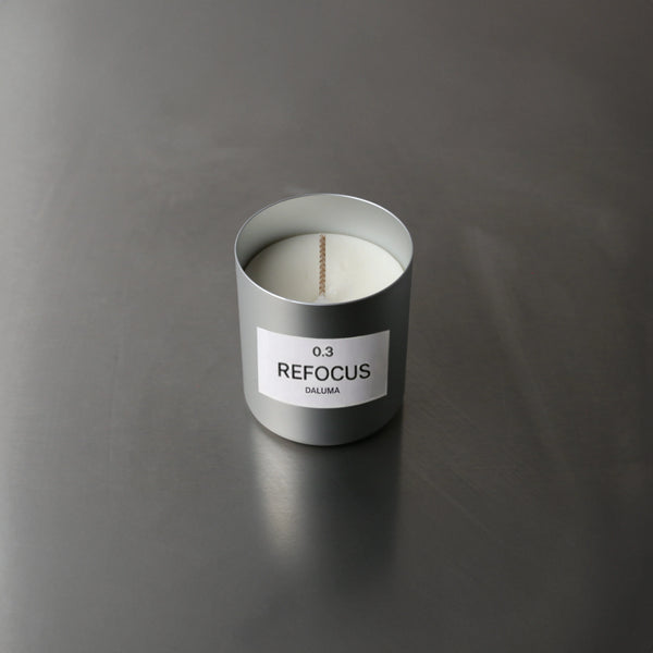 Ein silberner Nullpunkt DUFTKERZE GRADE 0.3 REFOCUS mit einem einzelnen zentrierten Docht ruht unbeleuchtet auf einer glatten grauen Oberfläche - perfekt für geistige Klarheit.