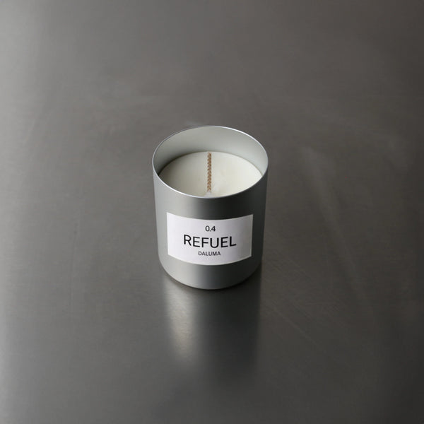 Ein silberner Nullpunkt-Kerzenhalter mit der Aufschrift DUFTKERZE GRADE 0.4 REFUEL ruht in der Draufsicht auf einer glatten grauen Fläche. Im Inneren bietet eine unbenutzte weiße Kerze einen Energie-Boost mit dem Duft von süßen Orangen - ideal für Duftkerzen-Fans.