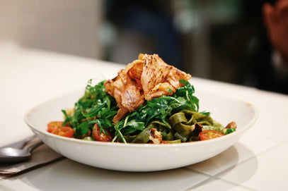 Eine weiße Schale mit grünen Nudeln, die mit gegrillten Hähnchenscheiben, Rucola, Kirschtomaten und Walnüssen belegt sind, steht auf einer weiß gekachelten Fläche neben einem Löffel und einer Serviette.