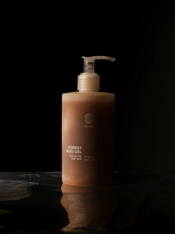 Eine hellbraune Pumpflasche mit der Aufschrift Forest Seed Gel steht vor einem schwarzen Hintergrund, der von sanftem Licht beleuchtet wird, mit subtilen Reflexen auf der Oberfläche darunter.