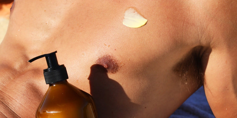 Eine Nahaufnahme der nackten Brust einer Person mit einem Klecks Sonnencreme neben der Brustwarze und einer braunen Pumpflasche in der Nähe, die das Auftragen von Sonnenschutzmitteln andeutet.