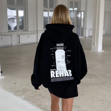 Eine blonde Person geht im Haus spazieren und trägt den DALUMA JOSEPH ATELIER X DALUMA REHAB HOODIE mit einer auffälligen Rückengrafik und dem Schriftzug "REHAB", gepaart mit weißen Sneakers. Der luftige Raum hat weiße Wände und große Fenster.