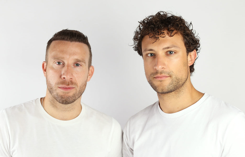 Zwei Männer mit kurzen Haaren, die schlichte weiße T-Shirts tragen, stehen nebeneinander vor einem weißen Hintergrund und blicken mit neutralem Gesichtsausdruck direkt in die Kamera.
