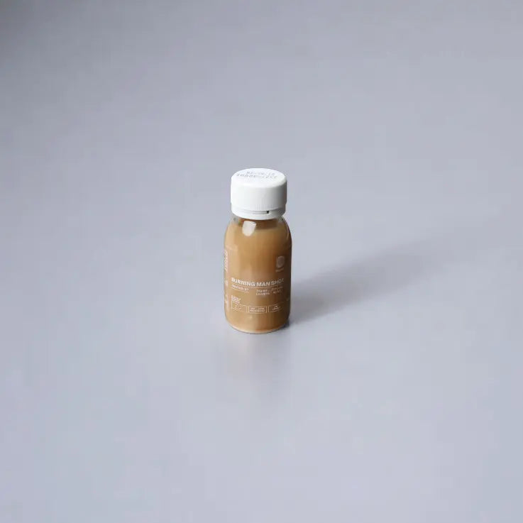 Eine kleine, mit hellbrauner Flüssigkeit gefüllte Flasche steht auf einer glatten, hellgrauen Oberfläche. Die Flasche hat einen weißen Deckel und ein Etikett mit weißer Schrift.