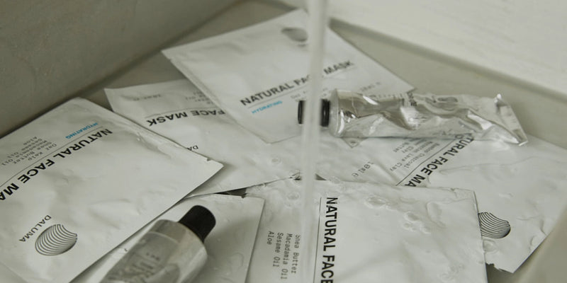 Mehrere weiße Päckchen mit der Aufschrift Natural Face Mask und Metalltuben liegen verstreut in einem Waschbecken unter fließendem Wasser, wobei auf den Päckchen und Tuben Tropfen zu sehen sind.