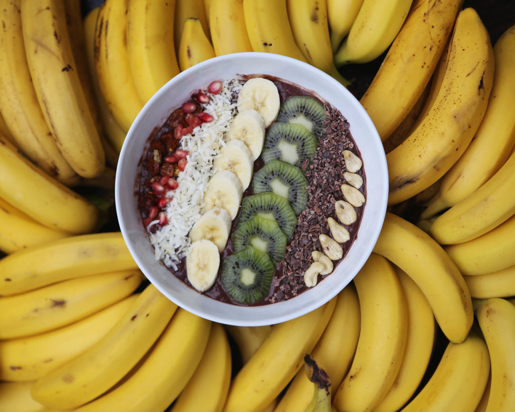 Eine Schale Smoothie mit Bananenscheiben, Kiwischeiben, Kokosraspeln, Granatapfelkernen, Kakaonibs und Nüssen, auf einem Bett aus reifen gelben Bananen.