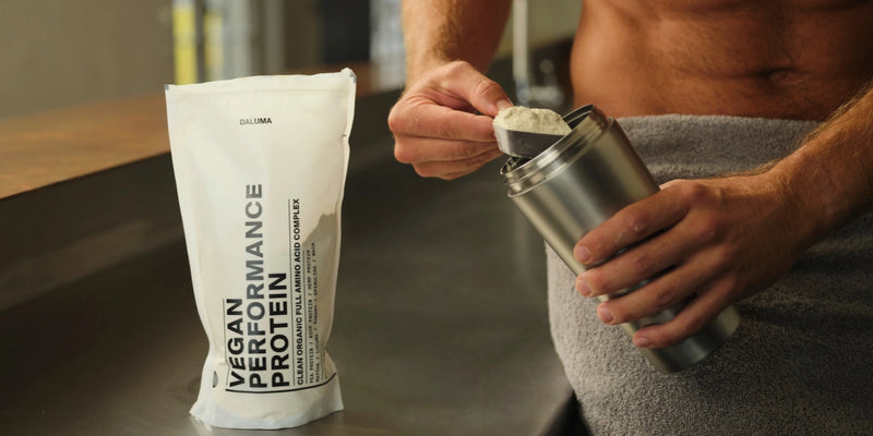 Eine in ein Handtuch gehüllte Person schöpft veganes Leistungsproteinpulver in einen Metallshaker. Der Beutel mit dem Proteinpulver steht aufrecht auf einer Arbeitsplatte neben ihnen.