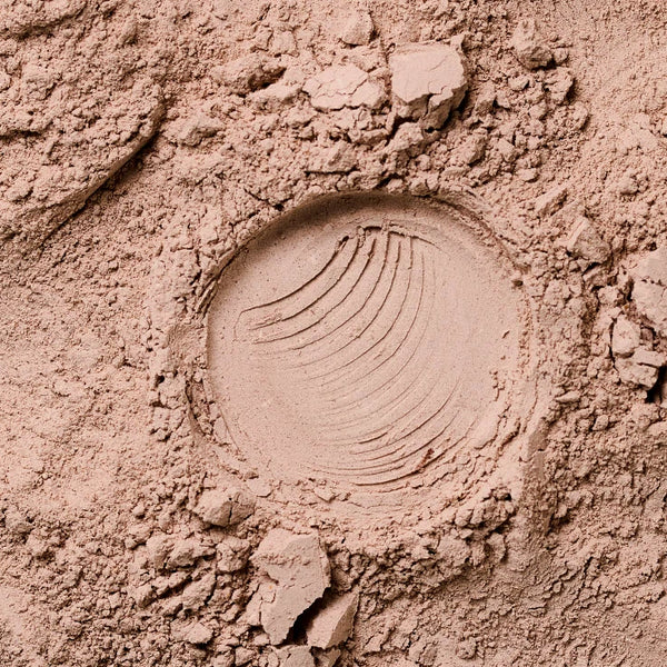 Nahaufnahme eines hellbraunen Pulvers mit einem kreisförmigen Abdruck in der Mitte, der geschwungene Linien zeigt und von kleinen Klumpen und losen Partikeln umgeben ist.