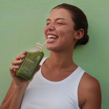 Eine lächelnde Frau in einem weißen Tank-Top hält eine Flasche grünen Saft und trinkt aus einem Strohhalm, während sie vor einer hellgrünen Wand steht.