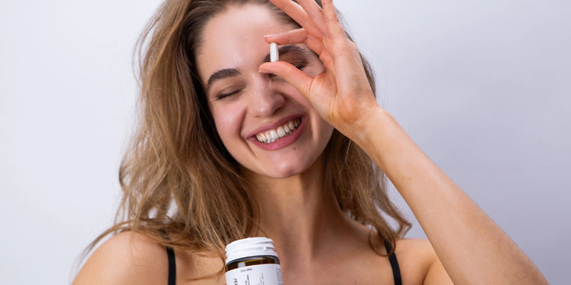 Eine lächelnde Frau mit langen Haaren hält eine offene Pillenflasche in der einen Hand und hält mit der anderen spielerisch eine Kapsel in die Nähe ihres Auges, vor einem schlichten hellen Hintergrund.