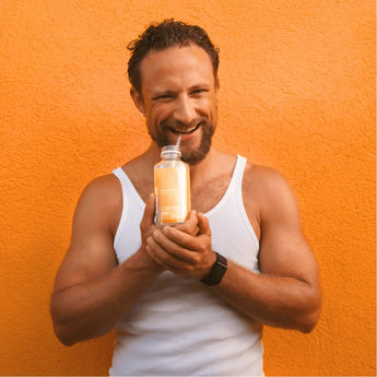 Ein lächelnder Mann in einem weißen Tank-Top hält eine Flasche mit orangefarbener Flüssigkeit in beiden Händen und steht vor einer leuchtend orangefarbenen, strukturierten Wand.