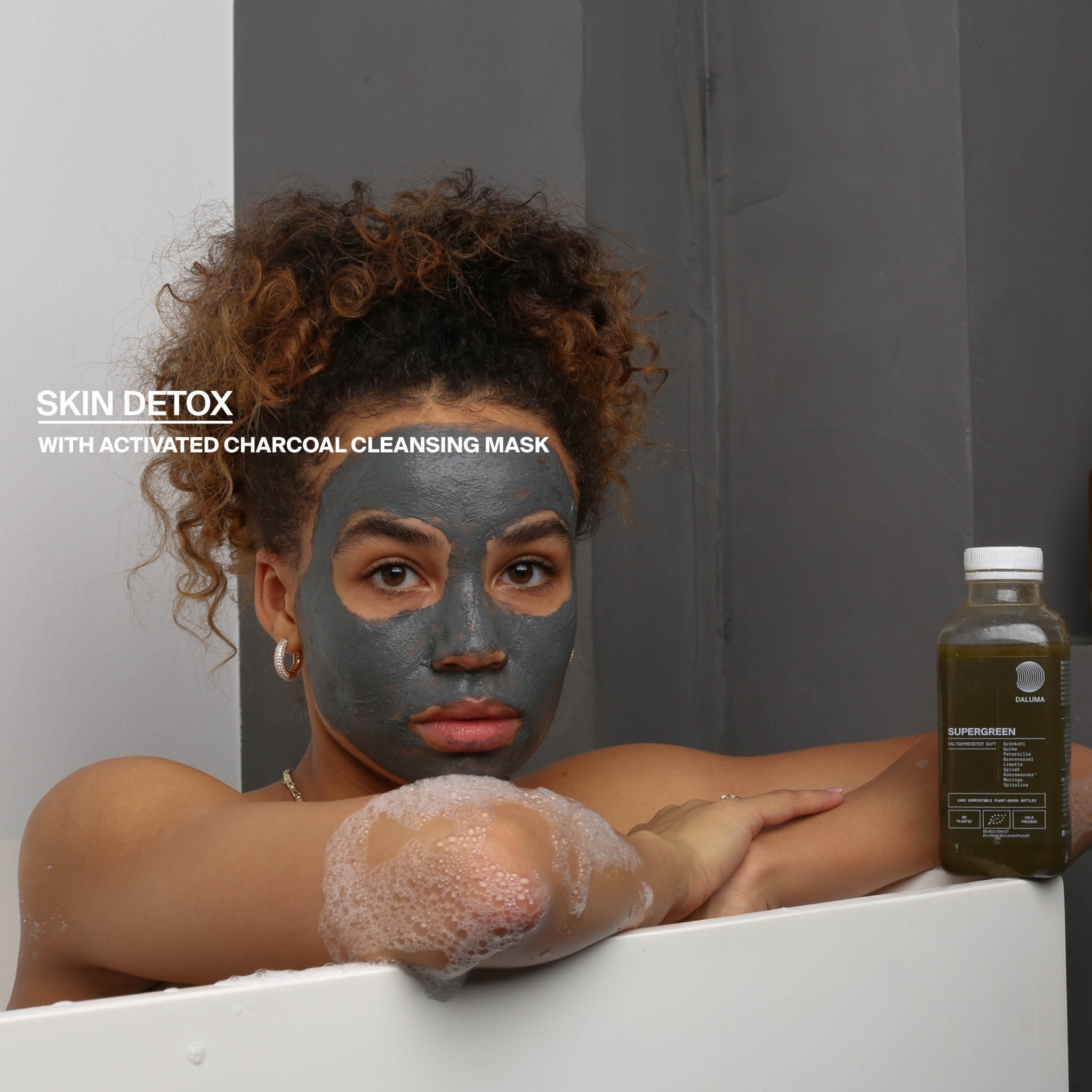 Eine Frau mit lockigem Haar entspannt sich in einem Schaumbad und trägt dabei eine Aktivkohle-Reinigungsmaske. Auf dem Wannenrand steht eine Flasche DALUMA BETTER ENERGY SAFTKUR 3 TAGE. Text: SKIN DETOX mit Aktivkohle-Reinigungsmaske.