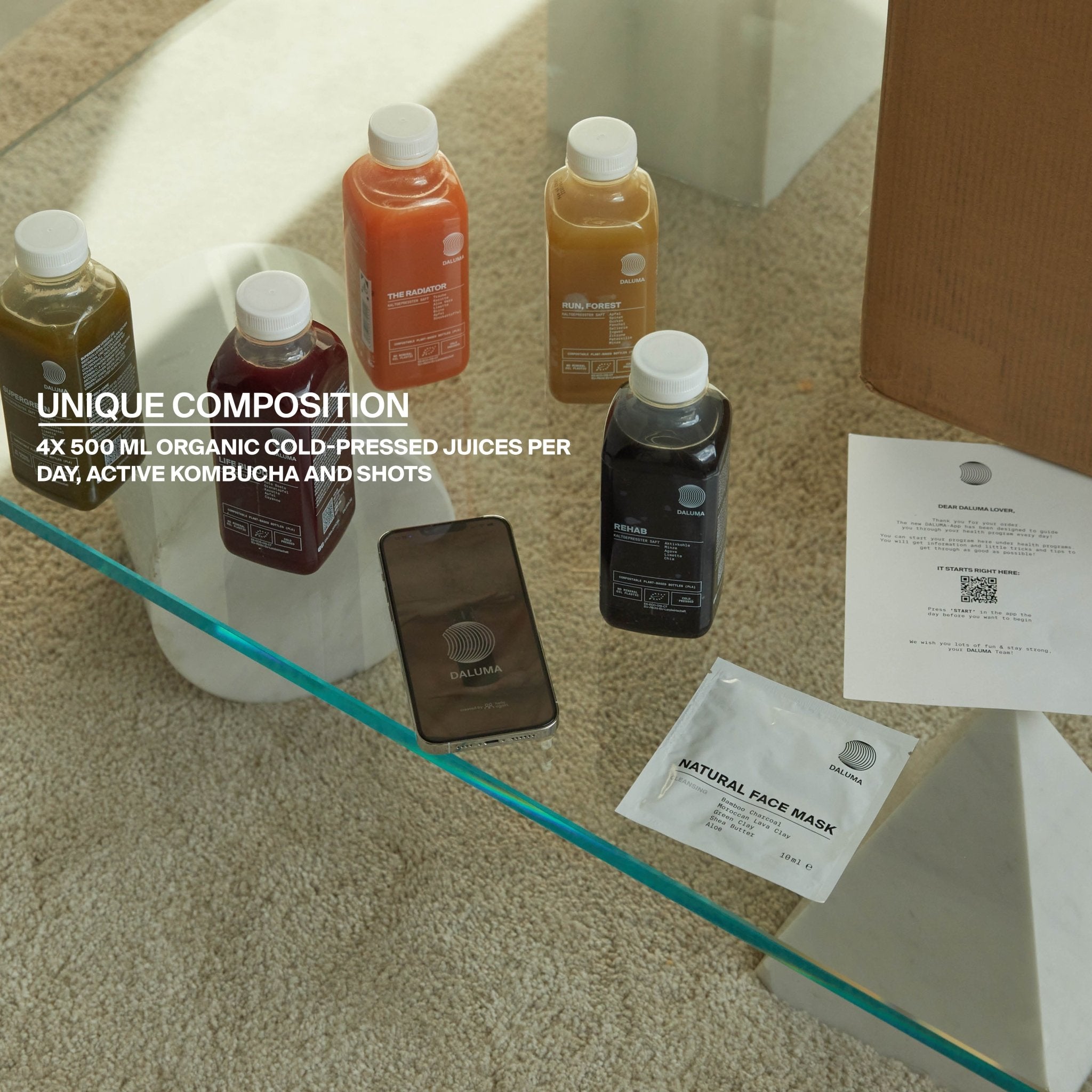 Auf einem Glastisch stehen mehrere Flaschen DALUMA BETTER ENERGY SAFTKUR 7 TAGE, ein Telefon, eine Packung Gesichtsmasken und eine Zeitung. Overlay: UNIQUE COMPOSITION 4x 500 ml Saftkur täglich, Aktiv Kombucha und Shots.