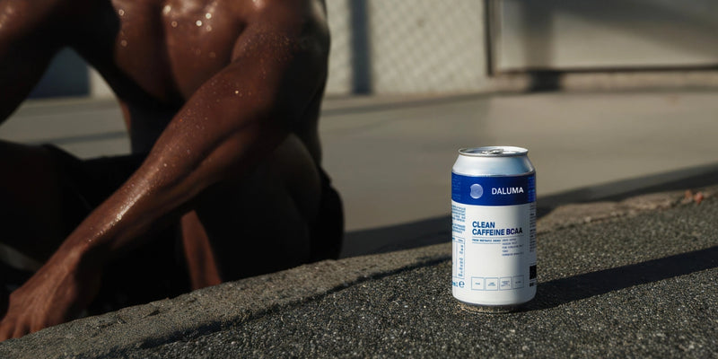 Eine Dose Daluma Clean Caffeine BCAA-Getränk liegt auf dem Bürgersteig, während im Hintergrund, teilweise unscharf, eine muskulöse Person mit verschwitzter Haut zu sehen ist.