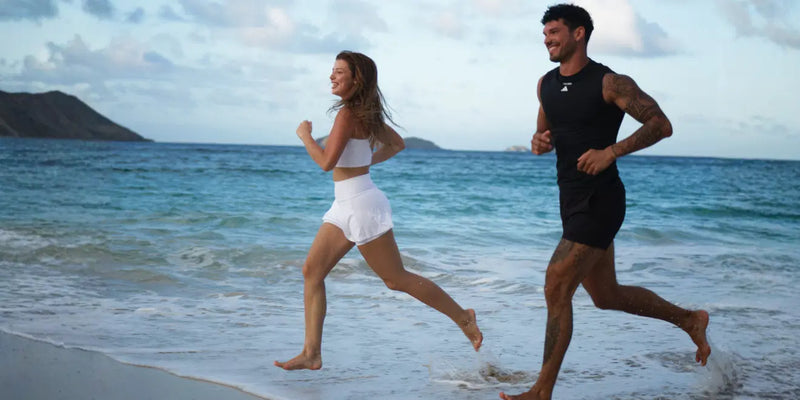 Eine Frau und ein Mann joggen am Ufer eines Sandstrandes entlang. Unter einem teilweise bewölkten Himmel sind die blauen Wellen des Meeres und die fernen Hügel zu sehen.