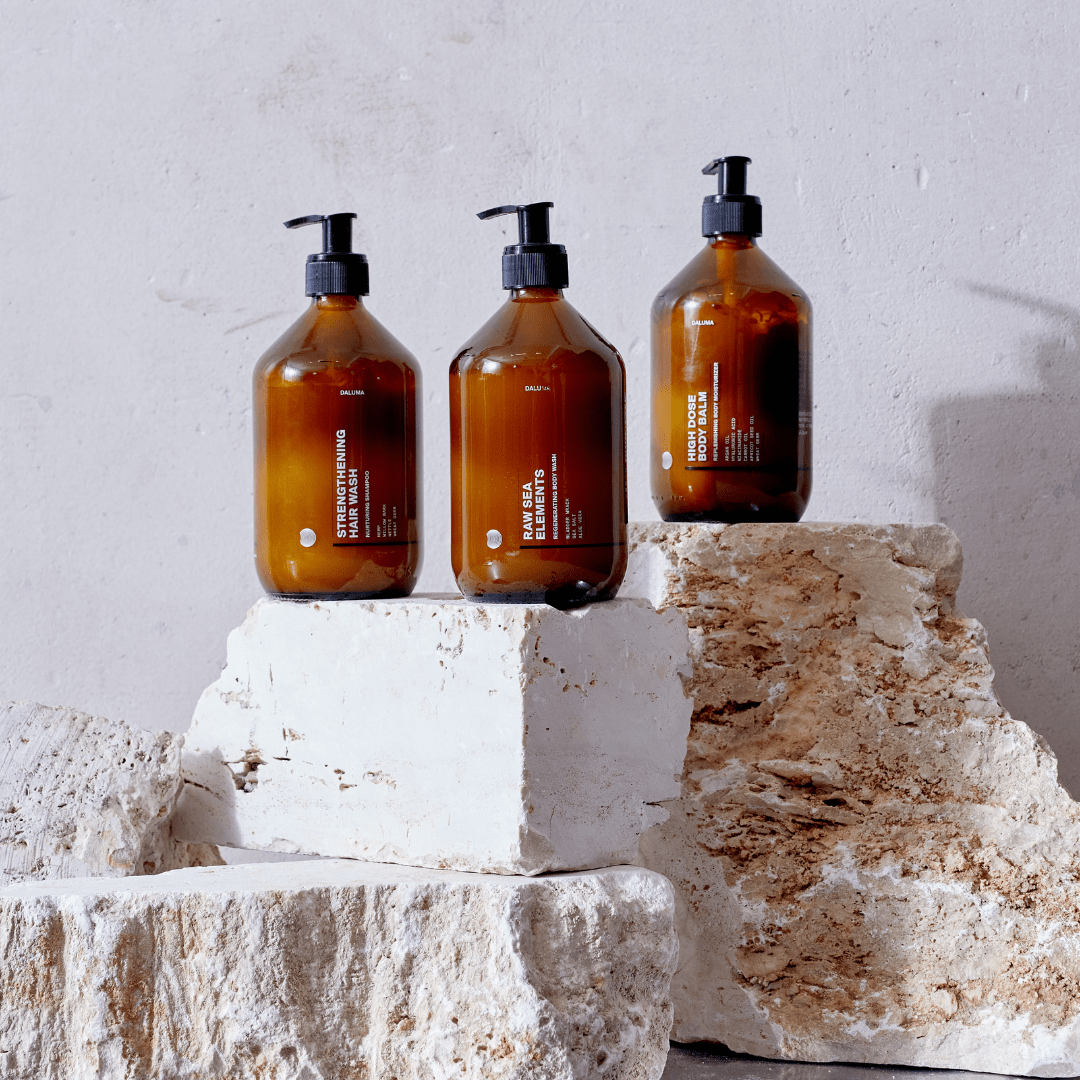 Drei bernsteinfarbene Pumpflaschen von DALUMA CLEAN CONDITIONER mit weißen Etiketten stehen auf strukturierten Steinblöcken vor einer hellgrauen Wand und präsentieren die pflegenden Formeln mit Süßmandelöl und Sheabutter.
