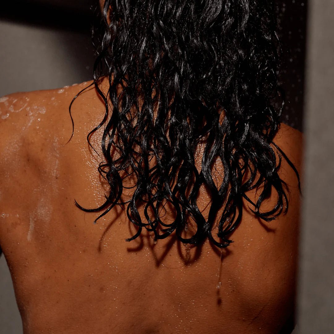 Eine Person mit nassem, lockigem, schwarzem Haar steht mit dem Rücken zur Kamera, mit Wassertropfen auf den nackten Schultern - ein Hinweis darauf, dass sie gerade mit DALUMA CLEAN CONDITIONER mit nährender Sheabutter geduscht hat.