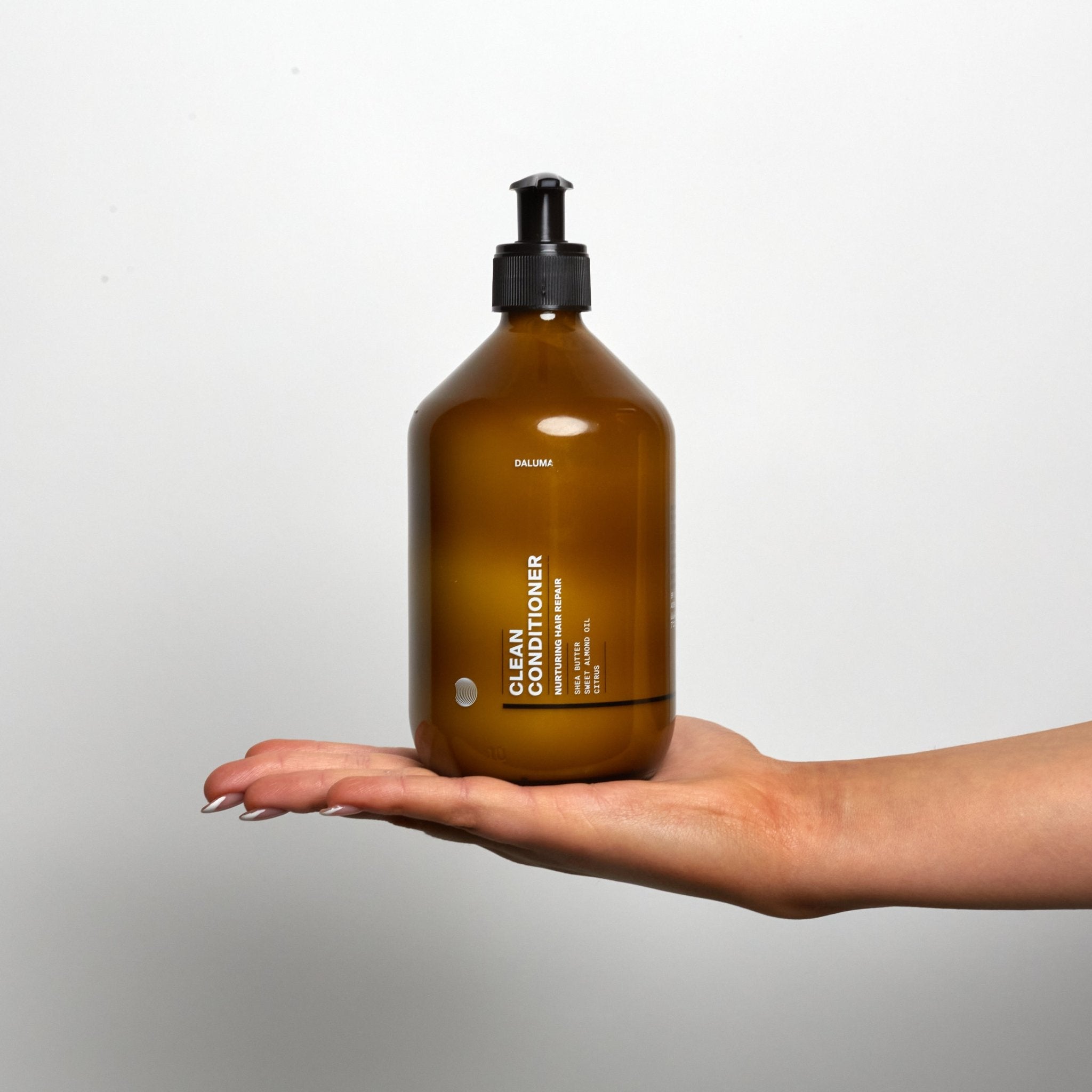 Eine Hand mit manikürten Nägeln hält eine braune Pumpflasche mit DALUMA CLEAN CONDITIONER, angereichert mit pflegendem Süßmandelöl, vor einem schlichten weißen Hintergrund.