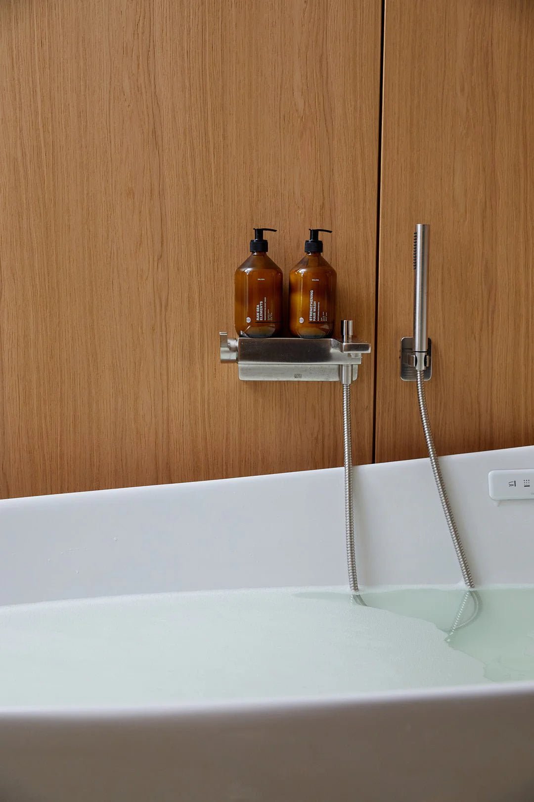Eine moderne, mit Wasser gefüllte Badewanne mit einem Metallregal, auf dem zwei bernsteinfarbene DALUMA CLEAN CONDITIONER-Flaschen stehen - eine davon mit Süßmandelöl angereichert - vor einer Holzwand mit einer eleganten Handbrause.