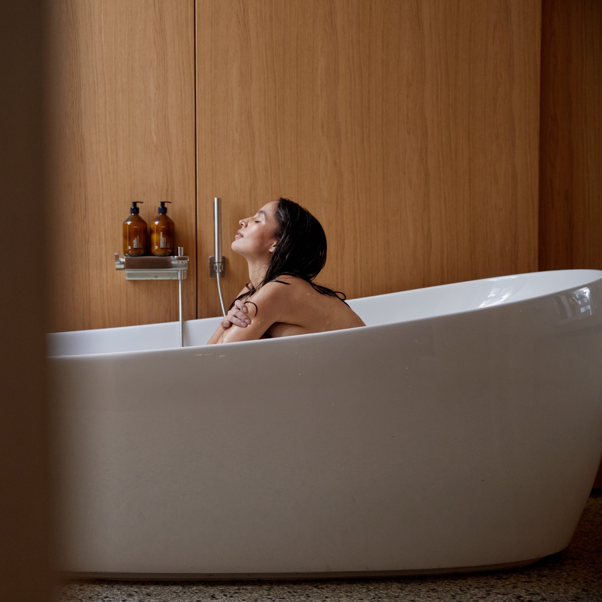 Eine Frau mit nassem Haar entspannt sich in einer weißen Badewanne, die Augen geschlossen und die Arme auf den Rand gestützt. Zwei Flaschen HAIR CARE DUO von Daluma-shop stehen in einem Regal, das an hölzernen Wandpaneelen befestigt ist.
