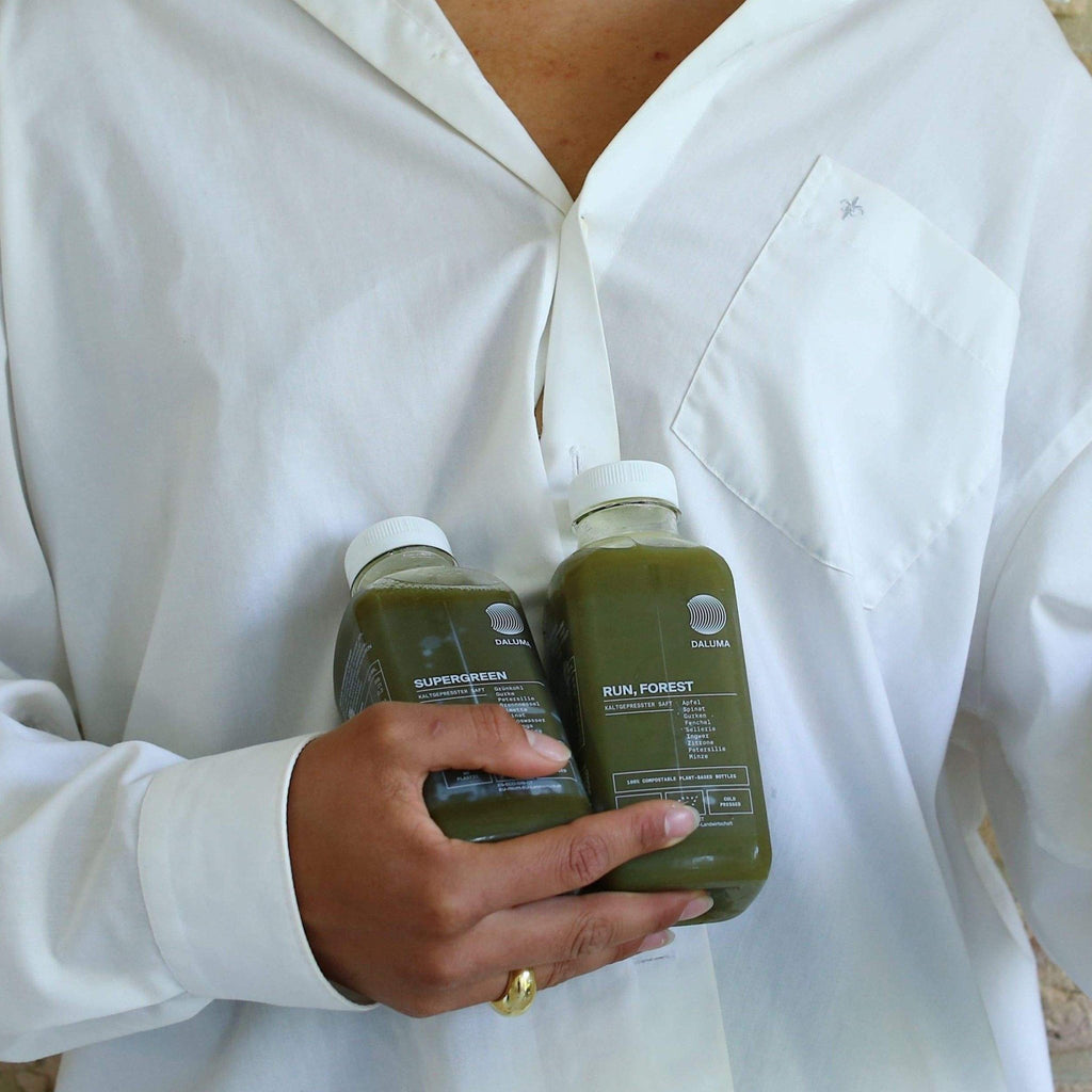 Eine Person in einem weißen Hemd hält zwei Flaschen des DALUMA DAILY GREEN JUICE PACKAGE mit den Aufschriften Super Forever und Run, Forest in der Hand. Die Flaschen sind mit weißen Kappen und minimalistischen Etiketten versehen - vollgepackt mit Mikronährstoffen für Ihren Daily Greens Boost.
