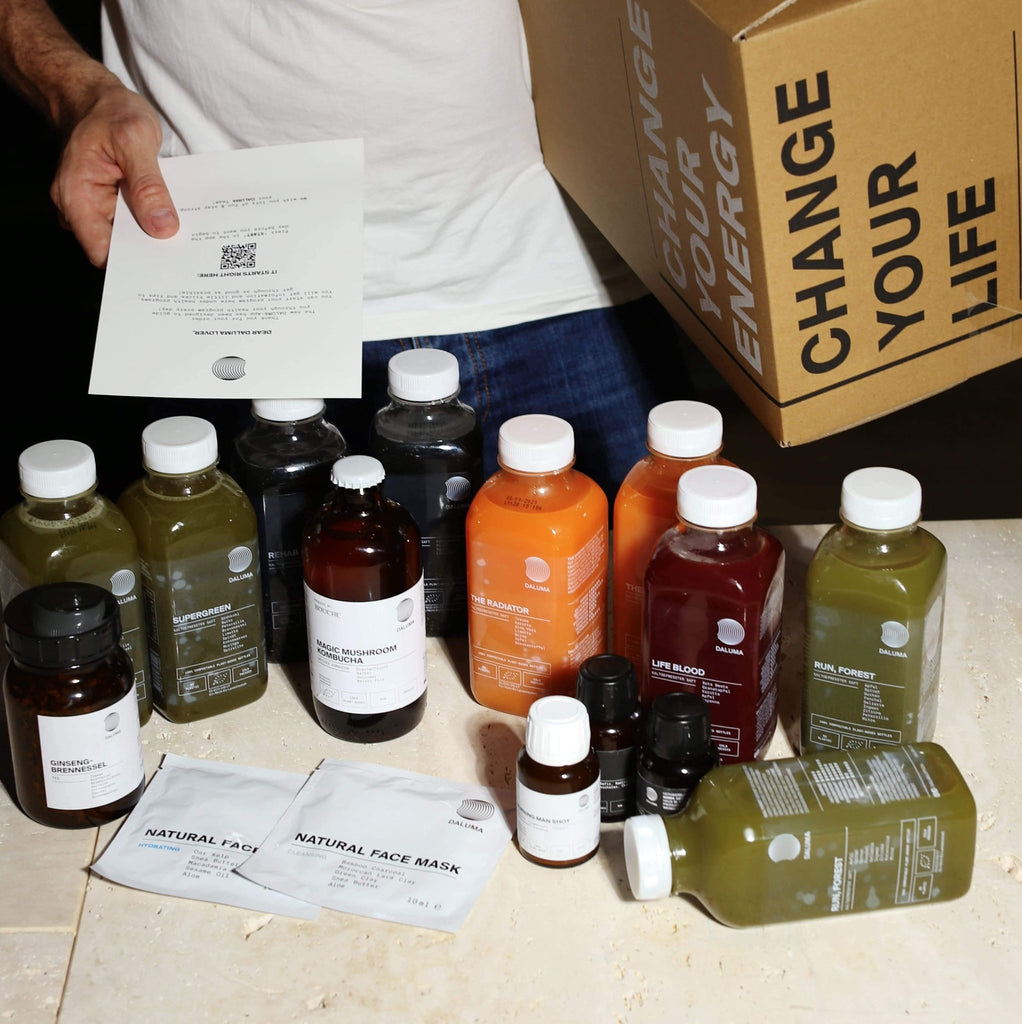 Eine Person hält eine Schachtel mit der Aufschrift DAILY HEALTH PACKAGE von DALUMA und ein Blatt Papier in der Hand, auf dem verschiedene Kaltpressverfahren Säfte, Vitamine und Gesichtsmasken für eine gesunde Routine auf einem beigen Tisch angeordnet sind.