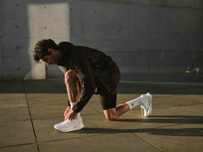 Ein Mann in sportlicher Kleidung kniet im Freien auf einer Betonfläche und bindet die Schnürsenkel seiner weißen Turnschuhe zu. Das Sonnenlicht wirft Schatten auf den Boden und im Hintergrund ist eine graue Wand zu sehen.