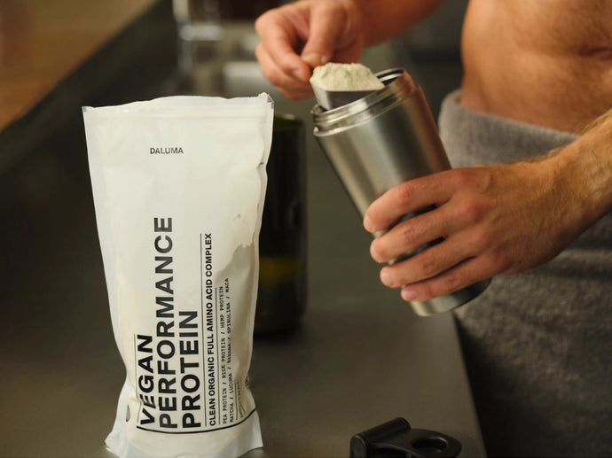 Eine Person schöpft Pulver aus einem Beutel mit der Aufschrift Vegan Performance Protein in eine Metallflasche mit Shaker auf einer Küchenzeile. Eine Schere liegt in der Nähe. Der Oberkörper der Person ist zu sehen, aber ihr Gesicht ist nicht abgebildet.