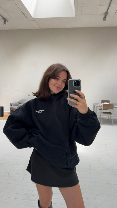 Eine Frau mit schulterlangen braunen Haaren macht ein Selfie im Spiegel in einem minimalistischen Raum. Sie trägt den DALUMA JOSEPH ATELIER X DALUMA REHAB HOODIE in Schwarz. Sie lächelt leicht. Unter einem Oberlicht sind Stühle und ein Sofa zu sehen.