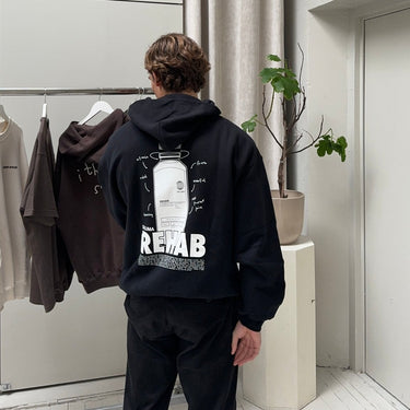 Eine Person mit kurzen braunen Haaren trägt den DALUMA JOSEPH ATELIER X DALUMA REHAB HOODIE mit einer Grafik und dem Text "REHAB" auf dem Rücken. Sie steht in einem minimalistischen Raum in der Nähe von aufgehängter Kleidung und einer Topfpflanze.