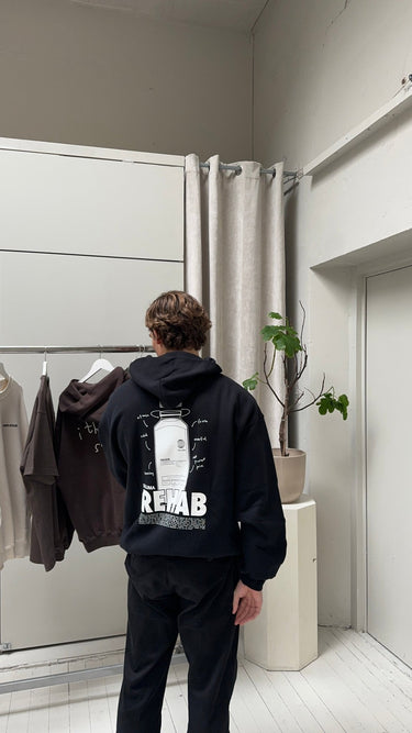 Eine Person mit gewellten braunen Haaren trägt den DALUMA JOSEPH ATELIER X DALUMA REHAB HOODIE - einen schwarzen Kapuzenpulli in Übergröße mit weißer Grafik und dem Schriftzug REHAB" auf dem Rücken - während sie in einem Innenraum neben einem Kleiderständer und einer Topfpflanze steht.