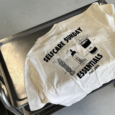 Das JOSEPH ATELIER X DALUMA SELFCARE SUNDAY TEE von DALUMA, ein T-Shirt aus nachhaltiger Bio-Baumwolle mit Oversize-Passform, trägt die Aufschrift "SELF-CARE SUNDAY ESSENTIALS DALUMA" und zeigt in schwarzer Schrift eine Kerze, eine Pflanze, ein Buch und ein Getränk.