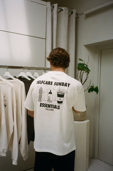 Eine Person mit kurzen, gewellten Haaren ist von hinten zu sehen, die das DALUMA JOSEPH ATELIER X DALUMA SELFCARE SUNDAY TEE trägt, ein übergroßes weißes T-Shirt mit schwarzen Abbildungen und Text. Sie stehen neben einem Kleiderständer und einer kleinen Topfpflanze.