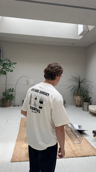 In einem hellen, minimalistischen Raum mit Pflanzen trägt eine Person mit kurzen braunen Haaren das DALUMA JOSEPH ATELIER X DALUMA SELFCARE SUNDAY TEE - ein weißes T-Shirt aus Bio-Baumwolle in Übergröße mit Selfcare Sunday Essentials und Illustrationen auf dem Rücken.