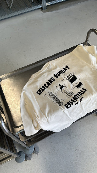 Das JOSEPH ATELIER X DALUMA SELFCARE SUNDAY TEE von DALUMA, ein übergroßes weißes Shirt aus Bio-Baumwolle, ruht auf einem Metallwagen. Es zeigt umweltfreundliche Siebdrucke von Selbstpflegeprodukten und den Text "SELF-CARE SUNDAY ESSENTIALS DALUMA".