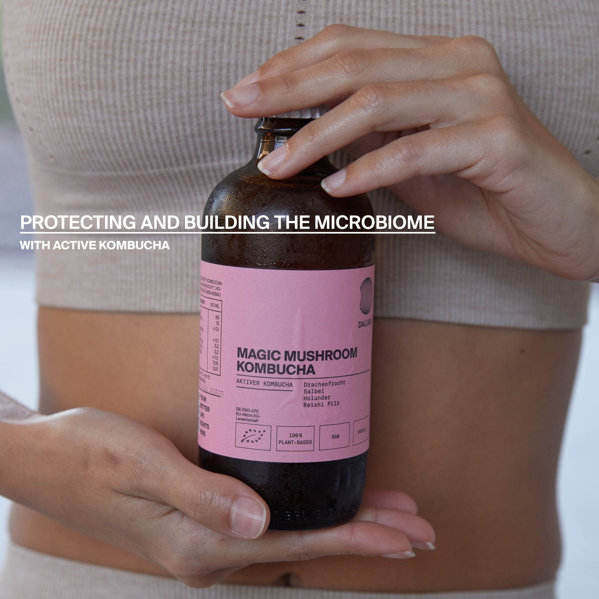 Eine Person in beiger Sportkleidung hält eine braune Flasche mit der Aufschrift CLEANSE DAY von DALUMA. Text überlagert: Unterstützen Sie Ihr Mikrobiom mit aktiver Kombucha - ideal für Ihre Saftkur oder bio-kaltgepresste Säfte-Routine.