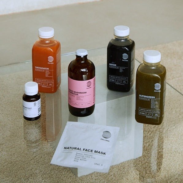 Fünf Flaschen DALUMA CLEANSE DAY und ein Beutel Natural Face Mask sind auf einem Glastisch angeordnet. Auf den Flaschen befinden sich verschiedenfarbige Flüssigkeiten, darunter auch Kombucha, die jeweils mit der Marke DALUMA beschriftet sind.