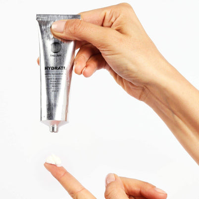 Eine Hand hält eine silberne Tube DALUMA FACIAL MOISTURIZER GESICHTSPFLEGE, während eine andere eine kleine Menge auf einem Zeigefinger sammelt, geeignet für alle Hauttypen, vor einem weißen Hintergrund.