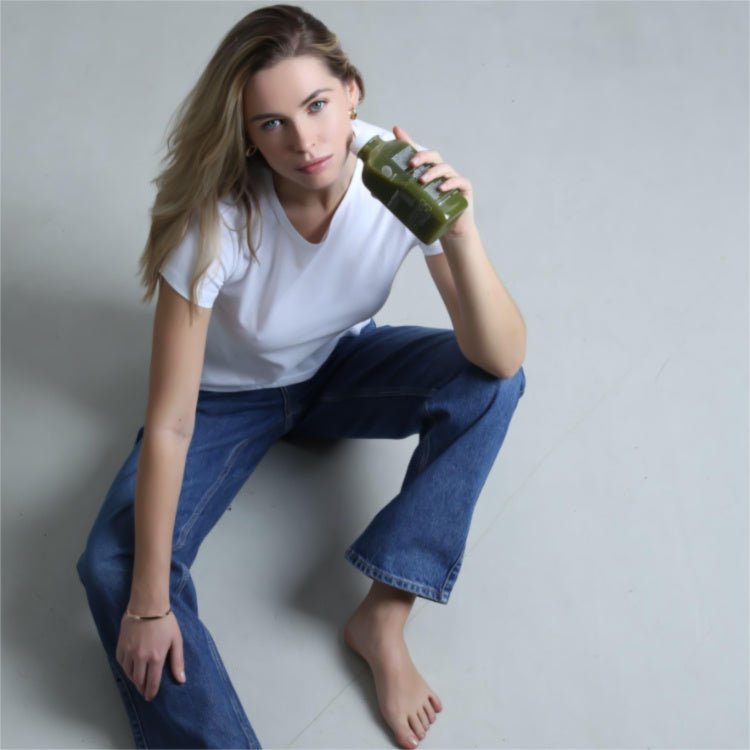 Eine Frau mit langen Haaren, weißem T-Shirt und blauer Jeans sitzt barfuß auf dem Boden, hält ein Glas GRÜNE SAFTKUR - 3 TAGE von DALUMA in der Hand und blickt in die Kamera.