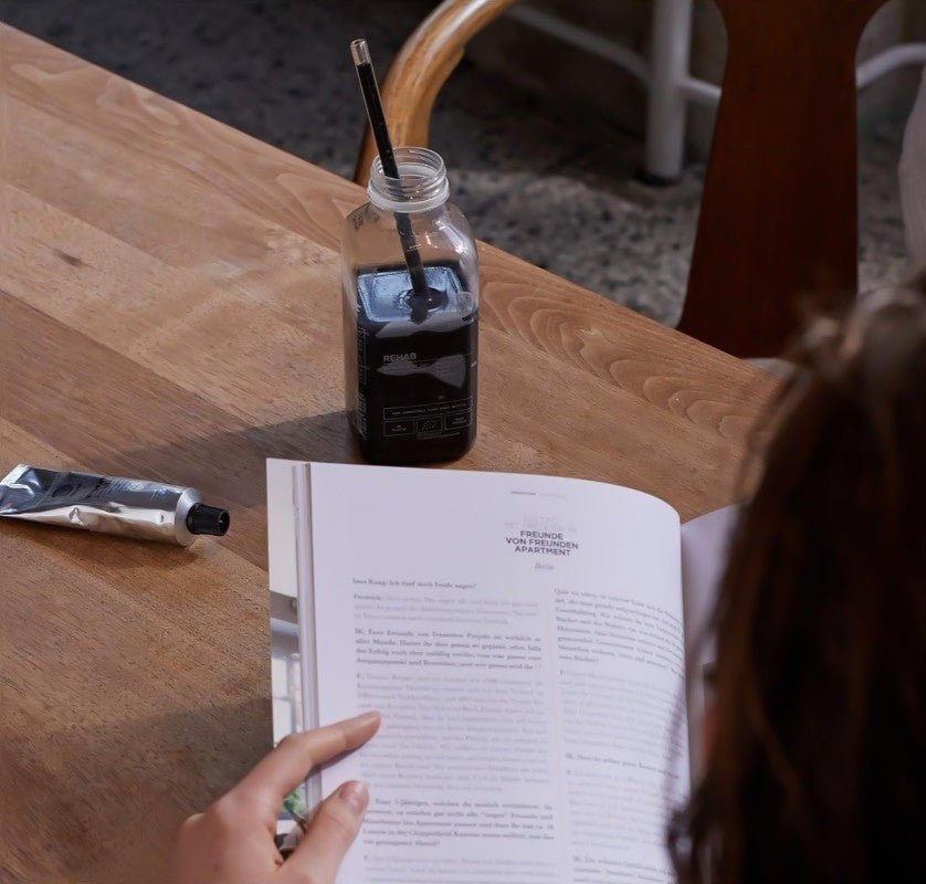 Eine Person liest ein Buch an einem Holztisch mit einer Flasche DALUMA GRÜNE SAFTKUR - 3 TAGE Saft, die einen Pinsel hält, und einer Tube Farbe neben sich.